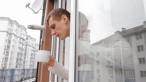 Young man looks out the window into the street and takes a sip of coffee 動画素材 127708372
