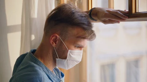 Young man looks out window and has medical mask on face during quarantine at Stock Footage 130142526