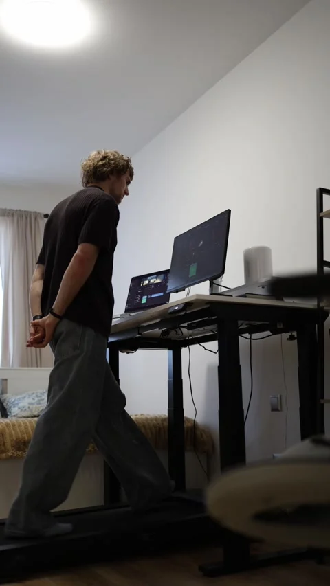 Young man lowers adjustable standing desk and continues walking on treadmill at  Stock Footage 346880728