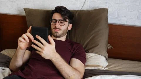 Young man lying in bed and reading ebook Stock Photos