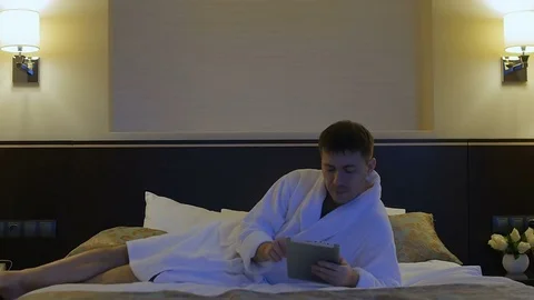 A young man is lying on a bed in a hotel and looks into the phone, tablet Stock Footage