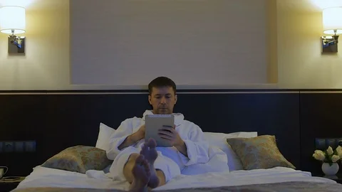 A young man is lying on a bed in a hotel and looks into the phone, tablet Stock-Footage 76117922