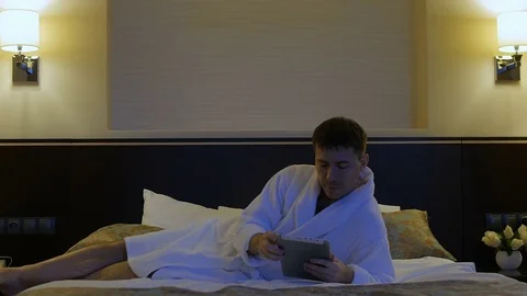 A young man is lying on a bed in a hotel and looks into the phone, tablet Stock-Footage 76118236