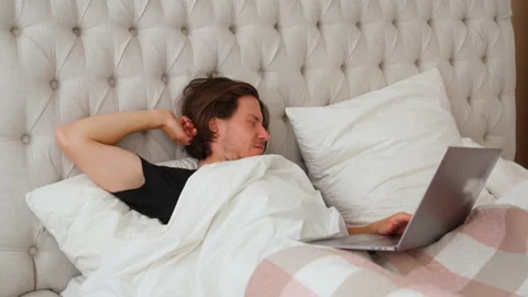 Young man lying in the bed with laptop make morning stretching and start working Stock Footage 138115385