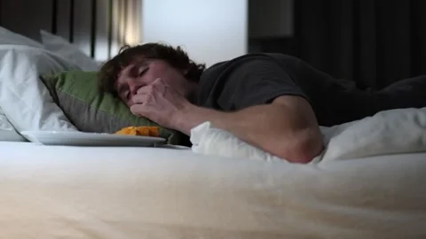 Young man lying in bed late at night reaching for food on a plate unhealthy life 库存影片 318217758
