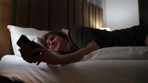 Young man lying in bed late at night watching series on smartphone illuminated b Stock Footage 318778732