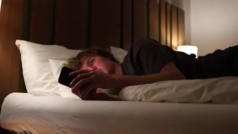 Young man lying in bed at night playing games on smartphone illuminated by warm  Stock Footage 318778720