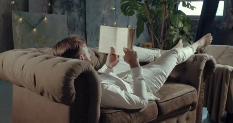 A young man lying on the couch reading a book in the apartment. Stock Footage 144502944