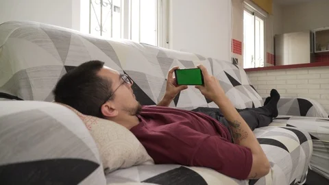 Young man lying on the couch staring at his cell phone, green screen Stock-Footage 129560655