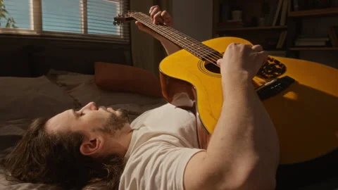 Young Man Lying down and Playing Acoustic Guitar at Home Stock Footage 337504985