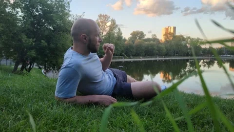 Young man lying on grass in public city park near pond Stockbeeldmateriaal 204159160
