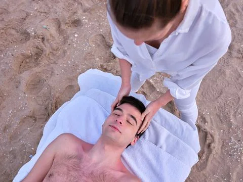 A young man lying on a massage table receiving a head massage on a beach in Stock Photos