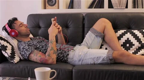 Young man lying on sofa and  listening to music on smartphone with headphone Stock Footage 51430943