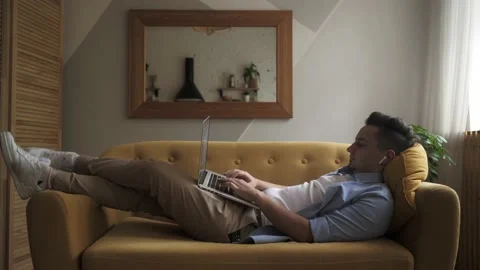 A young man is lying on the sofa with a computer on his lap and working Stock Footage 139628305