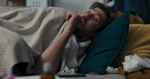 Young man is lying on sofa, covered with blanket. Brunette coughs a lot, he 스톡 동영상 218330185
