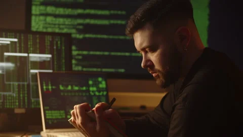 A young man makes notes in a notebook, sitting in front of monitors on which Stockbeeldmateriaal 236644414