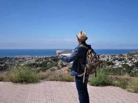 A young man with a map in his hands on the mountain looks at the village unde Stock Photos
