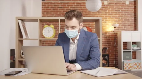 A young man in a mask sits at a table and types something into a laptop Stockbeeldmateriaal 188777826