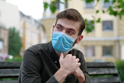 Young man in medical protective mask sits on the bench in city park Stock-Fotos