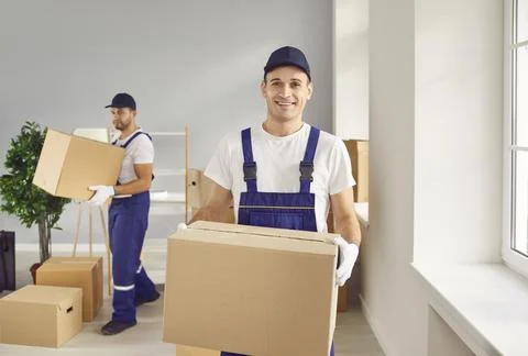 Young man mover worker in uniform holding a cardboard box and looking at camera Stock Photos