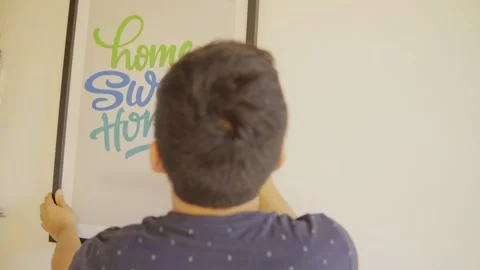 Young man moving into a new house, hanging a picture that says “Home sweet home. Vidéo 320405504