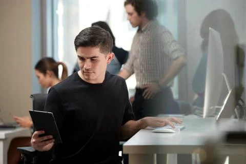 Young man multi-tasking by sitting at computer in office and checking tablet, Foto stock