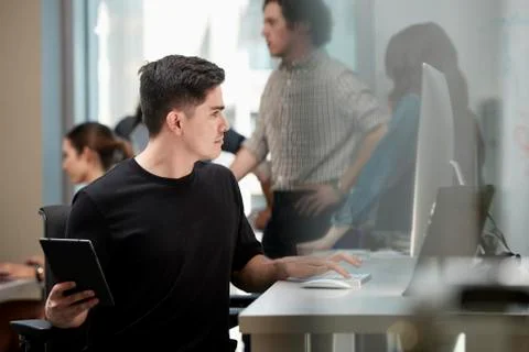 Young man multi-tasking by sitting at computer in office and checking tablet, Foto stock