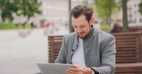 Young man with mustaches and a beard sitting on a bench in the square, typing on Stock Footage 140234953