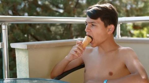 A young man with a naked torso eats ice cream while sitting at a glass table on Stock Footage 100219385