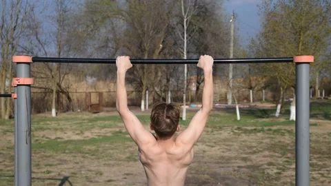 A young man with a naked torso pulls up on a horizontal bar on a sports field Stock Footage 131142893