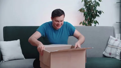 Young man opens a cardboard box while sitting on a home office couch. Stock Footage 145090667