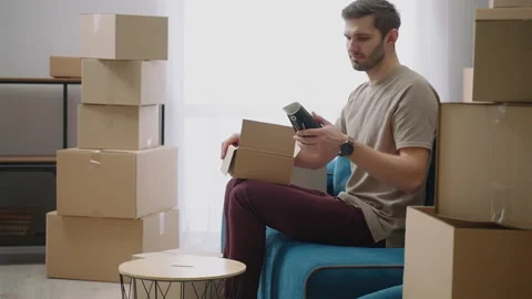 A young man opens a cardboard box and takes out things sitting on the sofa in a Stock Footage 149323442