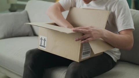 A young man opens a cardboard parcel on the couch in his living room 스톡 동영상 108549109
