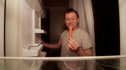 A young man opens the refrigerator at night and takes a sausage satisfied Stock Footage 144018666