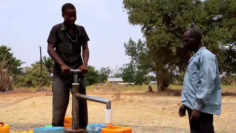 A young man operates a hand pump to collect water in yellow containers while an Vídeos de archivo 345835208