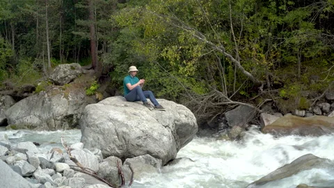 A young man in a Panama hat sits on a stone with a phone in his hands in the Stock Footage 140229288