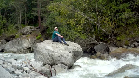 A young man in a Panama hat sits on a stone with headphones in the mountains by Stock Footage 140263179