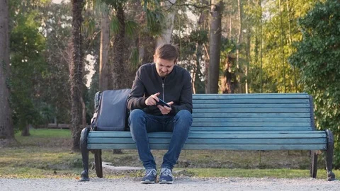 Young man in a Park on a bench is playing a game on his phone and Smoking Stock Footage 126239180