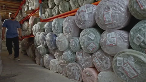 Young man passing by a stack of rolls of fabric lies in the warehouse of a Stock Footage 278962337
