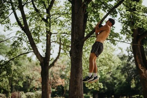Young man performing pull-ups on a tree branch in a park during an outdoor Stock Photos