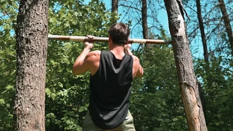 A young man performs pull-ups on a wooden bar set between two trees in a forest. Stockbeeldmateriaal 339016310