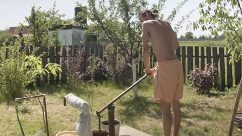 Young  man picking up cold water in a bucket from a column in his garden. 스톡 동영상 132580929