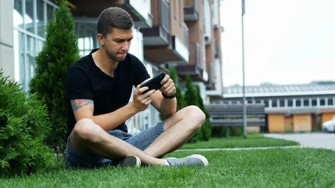Young man play mobile game on smartphone, sitting on the grass near the building Stock Footage 110817474