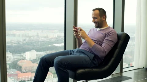 Young man playing game on smartphone while sitting on armchair with city view 스톡 동영상 66767967