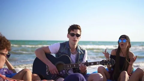 Young man playing guitar among group of friends sitting on easychairs on the Stock Footage 76717907