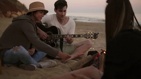 Young man is playing guitar by the fire sitting on the beach together with Stock Footage