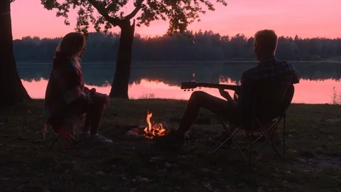 Young man is playing guitar to his girlfriend while sitting at bonfire at sunset Stock Footage 98852872