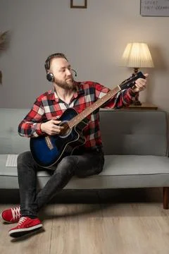Young man playing guitar while sitting on sofa in living room in the evening. Stock Photos