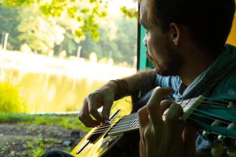Young man playing guitar while sitting in camping tent and looking away to river Stock Photos