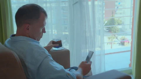 Young man pondering by the window with a cold drink and a phone in hand Stock Footage 282086170